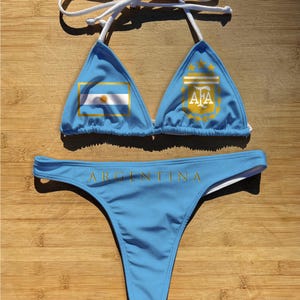 May include: Light blue bikini set featuring the Argentinian flag on the top and the word "ARGENTINA" on the bottom. The top has a triangle shape with white straps. The bottom is a thong style.