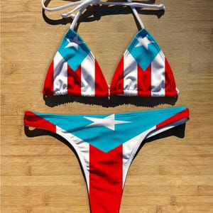 Könnte beinhalten: Ein zweiteiliges Bikini-Set mit der Flagge von Puerto Rico. Das Oberteil ist im Dreiecksstil mit weißen Bindebändern, und das Unterteil ist im String-Stil. Das Design beinhaltet rote, weiße und blaue Farben mit einem Stern.