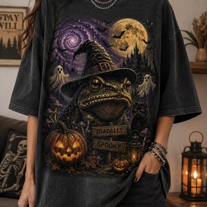 Op de afbeelding: Een antracietgrijs oversized T-shirt met een grafische print van een pad met een heksenhoed, een zwarte kat, een pompoen, geesten en een volle maan. De print bevat een houten bordje met de tekst TOADALLY SPOOKY in hoofdletters.
