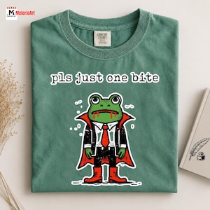 May include: A sage green t-shirt featuring a graphic of a green frog dressed as a vampire in a black suit with a red cape and red boots. The frog has red blood dripping from its mouth. Above the frog, the text reads: pls just one bite.