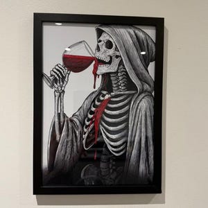 May include: Black framed art print featuring a skeletal figure in a hooded robe drinking red wine from a glass. The illustration is in black and white with red accents, depicting a macabre yet artistic scene. The artwork is mounted on a white background.