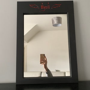 May include: A black framed mirror with a red devil design and wings at the top. The mirror is reflecting a person taking a selfie with a gold phone.
