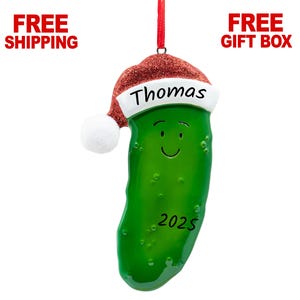 May include: Green pickle Christmas ornament with a red Santa hat and the name "Thomas" written on it. The ornament also has a white pom-pom and the year "2023" written on it.