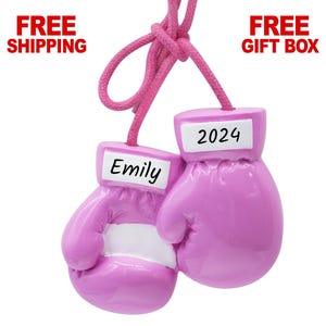 May include: A pair of pink boxing gloves ornament with white accents. One glove has the name "Emily" and the other has the year "2024" printed on it. The ornament is suspended by a pink cord.