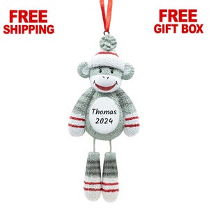 May include: A gray sock monkey ornament with a red ribbon for hanging. The monkey wears a white hat and has red accents on its arms and legs. The ornament has the name "Thomas" and the year "2024" on its belly.