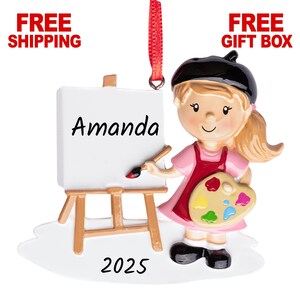May include: A figurine of a young artist wearing a pink dress and a black beret, holding a paintbrush and a palette. The artist stands next to a miniature easel with the name "Amanda" on the canvas and the year "2025" below. A red ribbon is attached for hanging.
