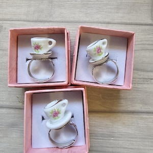 May include: Three silver-toned adjustable rings, each featuring a miniature white ceramic teacup and saucer ornament. The teacups are decorated with a small pink rose pattern and green leaves. Each ring is presented in an individual pink box.