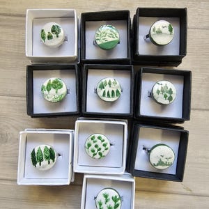 May include: A collection of ten rings, each featuring a circular top with hand-painted green botanical designs on a white background. The rings are individually presented in small, square black or white cardboard gift boxes with white inserts.