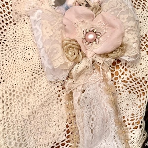May include: A decorative wall hanging featuring an oval portrait of a person in 18th-century attire, surrounded by layers of cream and white lace, fabric flowers in shades of pale pink and beige, and a central pink pearl-like bead accent.