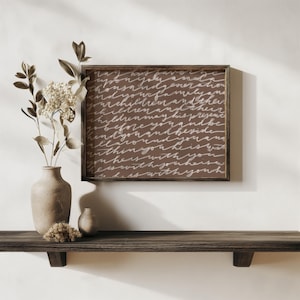 May include: A framed brown sign with white cursive text hangs above a wooden shelf. The sign is a square shape with a dark brown frame. A vase with dried flowers and a small vase are on the shelf.