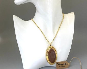 May include: A gold-toned necklace with a large, oval pendant featuring a brown center and gold border. The necklace is displayed on a white mannequin head. A small tag with the word "Vintage" is attached.