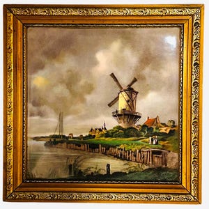 Op de afbeelding: Een ingelijst schilderij van een Nederlandse windmolen in een landelijk landschap. De windmolen is wit met een bruin dak en wordt omringd door groen gras en bomen. Op de voorgrond is een rivier met een klein bootje te zien. De lucht is bewolkt en grijs.