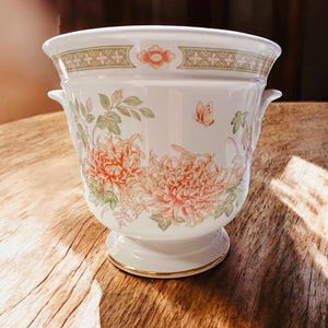May include: White ceramic planter with a floral design featuring large orange and pink flowers, green leaves, and a small butterfly. The planter has a gold rim and two small handles.