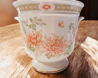 May include: White ceramic planter with a floral design featuring large orange and pink flowers, green leaves, and a small butterfly. The planter has a gold rim and two small handles.