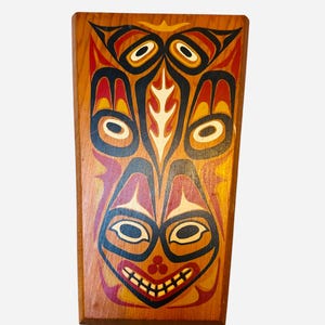 May include: A wooden plaque featuring a painted design of a stylised face with three eyes, a mouth, and a nose. The face is painted in red, orange, yellow, black, and white.