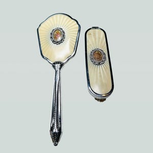 Könnte beinhalten: Ein Vintage-Kosmetikset mit einem Handspiegel und einer Bürste. Das Set hat ein cremefarbenes, fächerförmiges Design mit einem Blumenzentrum und silberfarbenen Metallbesatz. Der Spiegel hat einen langen, verzierten Griff.