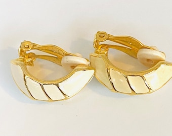 May include: A pair of gold tone clip-on earrings with white enamel detailing. The earrings are shaped like a half-circle and have a geometric design.