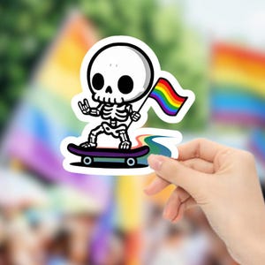May include: A sticker featuring a cartoon skeleton riding a skateboard and waving a rainbow flag. The skeleton is white with black eye sockets and a black nose. The skateboard is purple with black wheels. The background is blurred with rainbow colors.