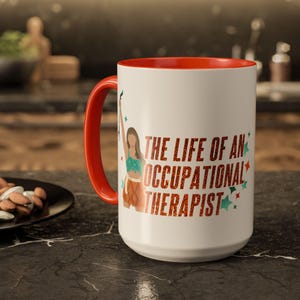 May include: White ceramic mug with a red interior and handle. The mug features the text "THE LIFE OF AN OCCUPATIONAL THERAPIST" in red and a graphic of a person. The mug is on a dark surface.