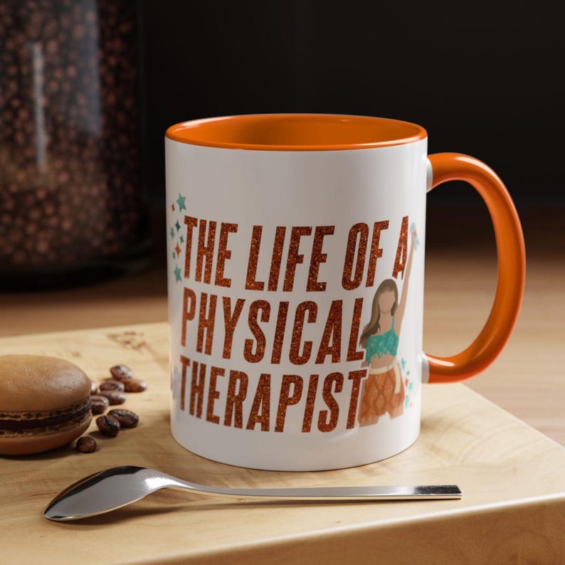 May include: White ceramic mug with an orange interior and handle. The mug features the text "THE LIFE OF A PHYSICAL THERAPIST" in orange lettering, along with a graphic of a person. A spoon and a pastry are on the wooden surface.