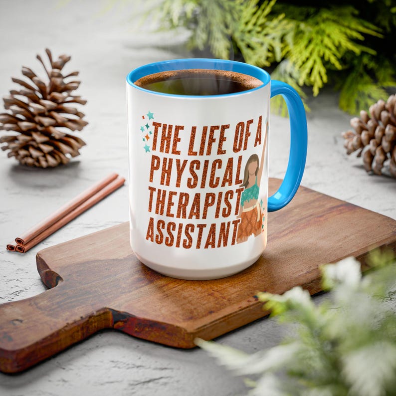 May include: White ceramic mug with a blue interior and handle, filled with coffee. The mug features the text "THE LIFE OF A PHYSICAL THERAPIST ASSISTANT" in orange lettering, along with a graphic of a person. The mug sits on a wooden cutting board.