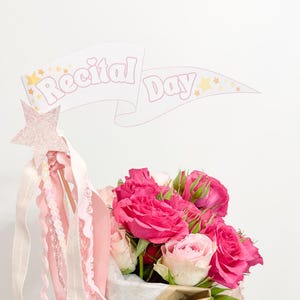 May include: A bouquet of vibrant hot pink and soft pale pink roses wrapped in white tissue paper and tied with a white tulle bow. A pink glittery star wand with dangling ribbons and a pennant flag featuring the text Recital Day are attached.