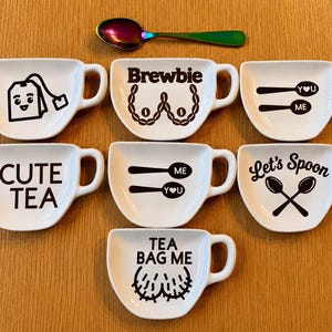 May include: Seven white ceramic tea bag holders with black graphic designs and text, including phrases like Cute Tea, Brewbie, and Let's Spoon, accompanied by a single iridescent rainbow-coloured metal spoon positioned at the top centre.