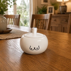 May include: A white ceramic sugar bowl with a lid and a black graphic of breasts. The bowl is round and sits on a wooden table. The lid has a small, round handle. The background includes a dining room table setting.