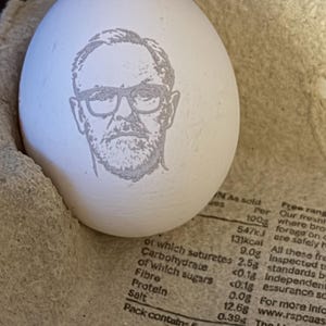May include: A white egg featuring a laser-engraved portrait of a person with a beard and glasses. The egg is positioned inside a textured, light brown cardboard carton that displays nutritional information and text regarding egg quality standards.
