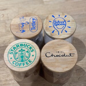 May include: Four small, cylindrical containers with wooden lids. The lids feature logos: Tetley tea, Silver Spoon, Starbucks Coffee, and Hotel Chocolat. The containers are made of a light-coloured wood and are arranged on a wooden surface.