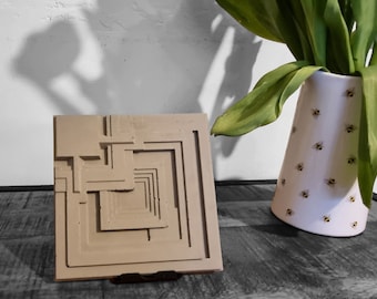 May include: A beige concrete tile with a geometric design, resembling a building facade, is displayed on a wooden surface. The tile is on a small black stand. A white vase with a bee pattern is in the background.