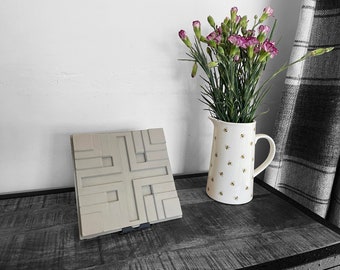 May include: A gray geometric design tile with a raised pattern. The tile is on a dark wood surface. A white pitcher with a bee pattern is filled with pink flowers.