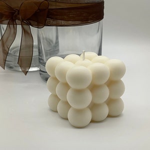 Peut inclure: Une bougie blanche ronde à bulles avec une seule mèche. La bougie est en forme de cube et a une surface texturée.