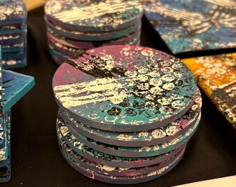 Op de afbeelding: Een stapel van zes ronde onderzetters met een abstract ontwerp in blauw, roze, wit en zwart. De onderzetters zijn gemaakt van hout en hebben een verweerde look.