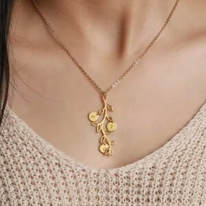 May include: A gold-colored necklace with a delicate branch pendant. The pendant features three circular charms, each engraved with a single initial. The necklace is worn against a cream-colored sweater.