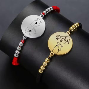 Puede incluir: Dos pulseras a juego con colgantes circulares con contornos de mapas y detalles de corazón. Una pulsera tiene un colgante plateado con un cordón rojo y cuentas plateadas. La otra tiene un colgante dorado con un cordón negro y cuentas doradas.