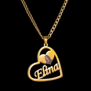 Pode incluir: Um colar dourado com um pingente em forma de coração. O pingente apresenta o nome "Elina" em fonte cursiva e uma pequena foto dentro de um coração. A corrente é do estilo elo de freio, em um fundo preto.
