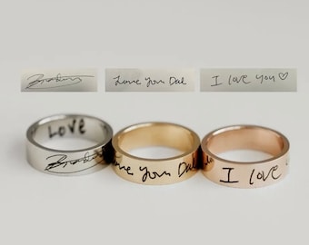 Puede incluir: Tres anillos de plata, oro y oro rosa con mensajes escritos a mano grabados en ellos. Los mensajes son "Love", "Love you Dad" e "I love you".