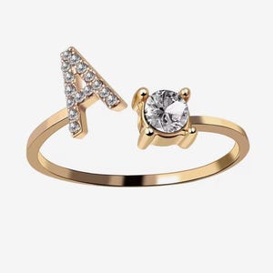 Peut inclure: Bague dorée avec la lettre "A" incrustée de petits pierres précieuses transparentes. La bague comporte également une grosse pierre précieuse ronde et transparente. La bague est posée sur un fond blanc uni.