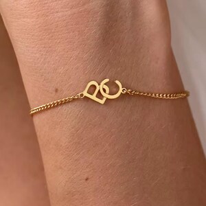 Könnte beinhalten: Ein goldfarbenes Armband mit einer filigranen Kette. Das Armband zeigt zwei verbundene Initialen, "B" und "C", in einer stilisierten Schriftart. Das Armband wird am Handgelenk einer Person getragen.