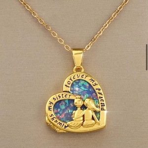 Peut inclure: Collier en or avec un pendentif en forme de cœur représentant deux personnages se tenant la main. Le pendentif est recouvert de paillettes irisées et les mots "Always my sister forever my friend" sont inscrits sur le bord.