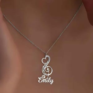 May include: A silver necklace with a treble clef pendant and the name "Emily" in a cursive font. The pendant features a heart-shaped design at the top of the clef. The necklace is worn around the neck.