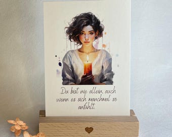 May include: A watercolor illustration of a woman holding a lit candle. The background is a soft watercolor wash of pink and blue. The text reads "Du bist nie allein, auch wenn es sich manchmal so anfühlt."