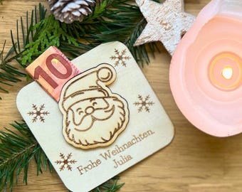 May include: A wooden Christmas gift card holder with a Santa Claus design and snowflake accents. A ten-euro bill is partially inserted. The card reads "Frohe Weihnachten Julia". A lit candle, star, and pinecone are in the background.