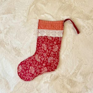May include: A quilted Christmas stocking with a red floral pattern, white lace trim, and a coral top. The stocking has a red loop for hanging.