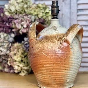 May include: A ceramic lamp base in cream and orange. The lamp features two handles and a ribbed texture. It has a black lamp socket. Dried flowers are in the background. The lamp is a decorative item.