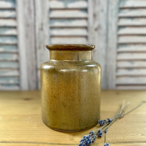 May include: An olive-green ceramic vase with a dark brown rim. A small bunch of dried lavender with purple flowers sits on a wooden surface beside the vase. The background shows a white wooden shutter. The vase is approximately 15 cm tall.