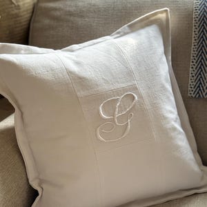 May include: A square, cream-coloured decorative cushion with a stitched, framed design. The cushion features a large, embroidered white letter "G" in the centre. The cushion is placed on a neutral-toned sofa.