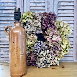 May include: A brown stoneware lamp base with a handle and a black lamp socket. Next to it is a wreath of dried hydrangeas in shades of purple, green, and cream. The background features a weathered, light blue shutter.