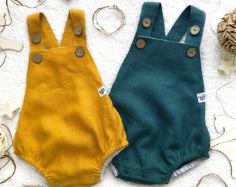 May include: Two baby rompers, one mustard yellow and one teal green, with adjustable straps and button closures. Both rompers have a small white label with the text "Smiley" on them.